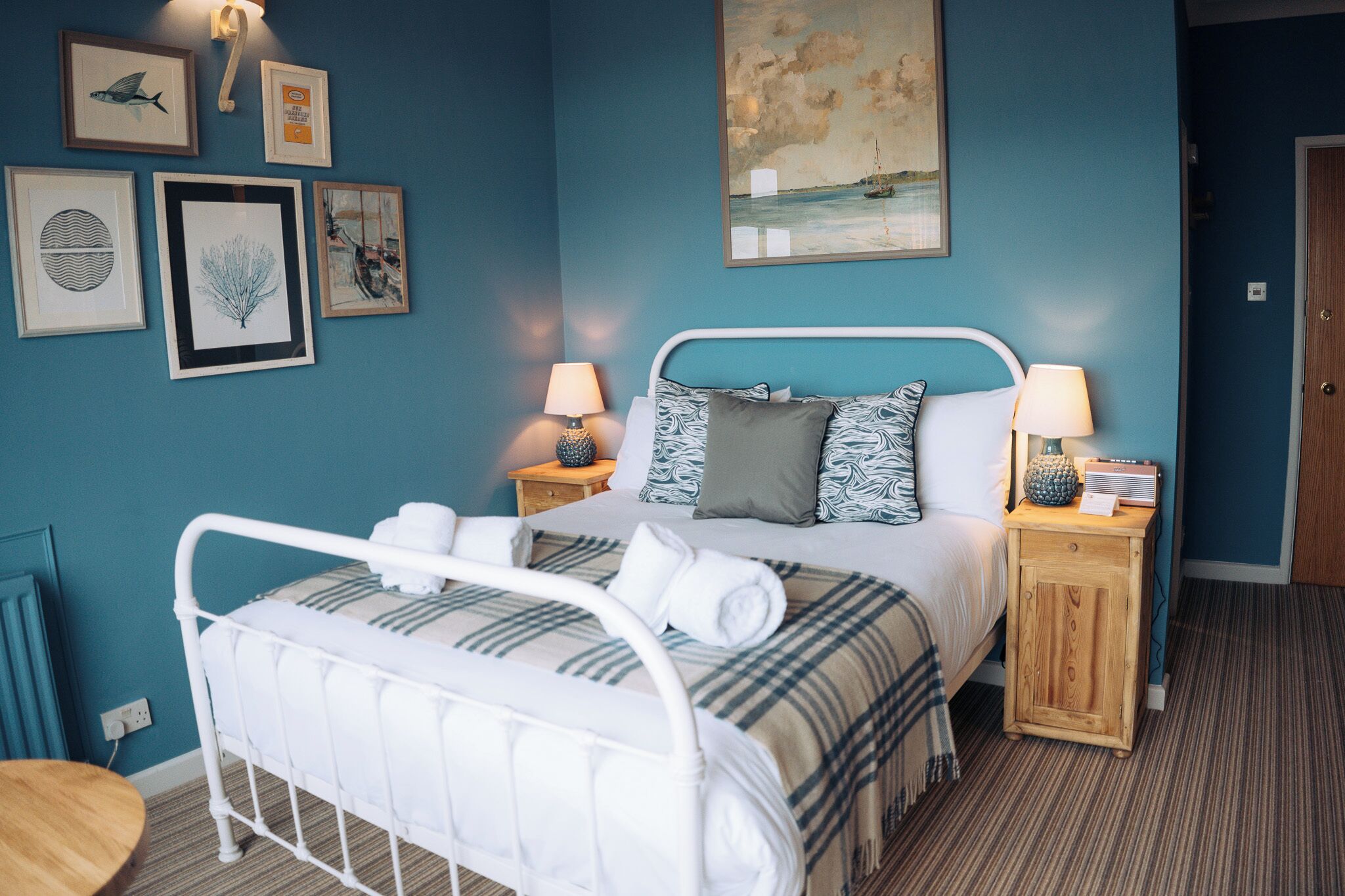 A bedroom with a bed, two bedside tables, a nightstand, a wall with framed artwork, and a door.