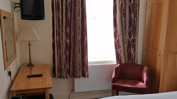 A hotel room with a bed, a desk with a lamp, a chair, a wardrobe, and a TV mounted on the wall.