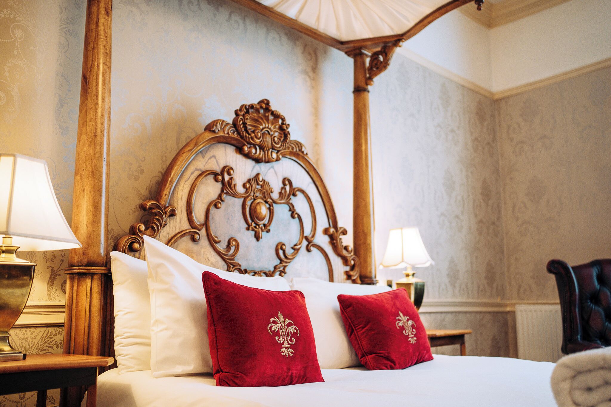 Una cama con dosel y tablero de cabecera ornamentado, sábanas blancas y almohadones rojos con bordados dorados.