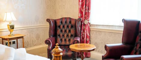 Una stanza con un letto, un comodino in legno con una lampada, una poltrona in pelle trapuntata, un tavolino rotondo in legno e una tenda rossa.