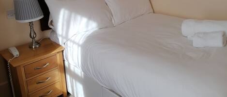 A neatly made bed with white linens, a wooden nightstand with a lamp, and a painting of flowers on the wall.