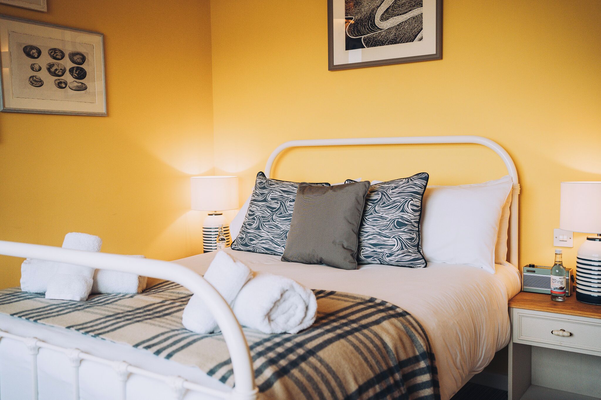 A hotel room with a bed, pillows, a bedside table with a lamp, a bottle of water, and a framed picture on the wall.