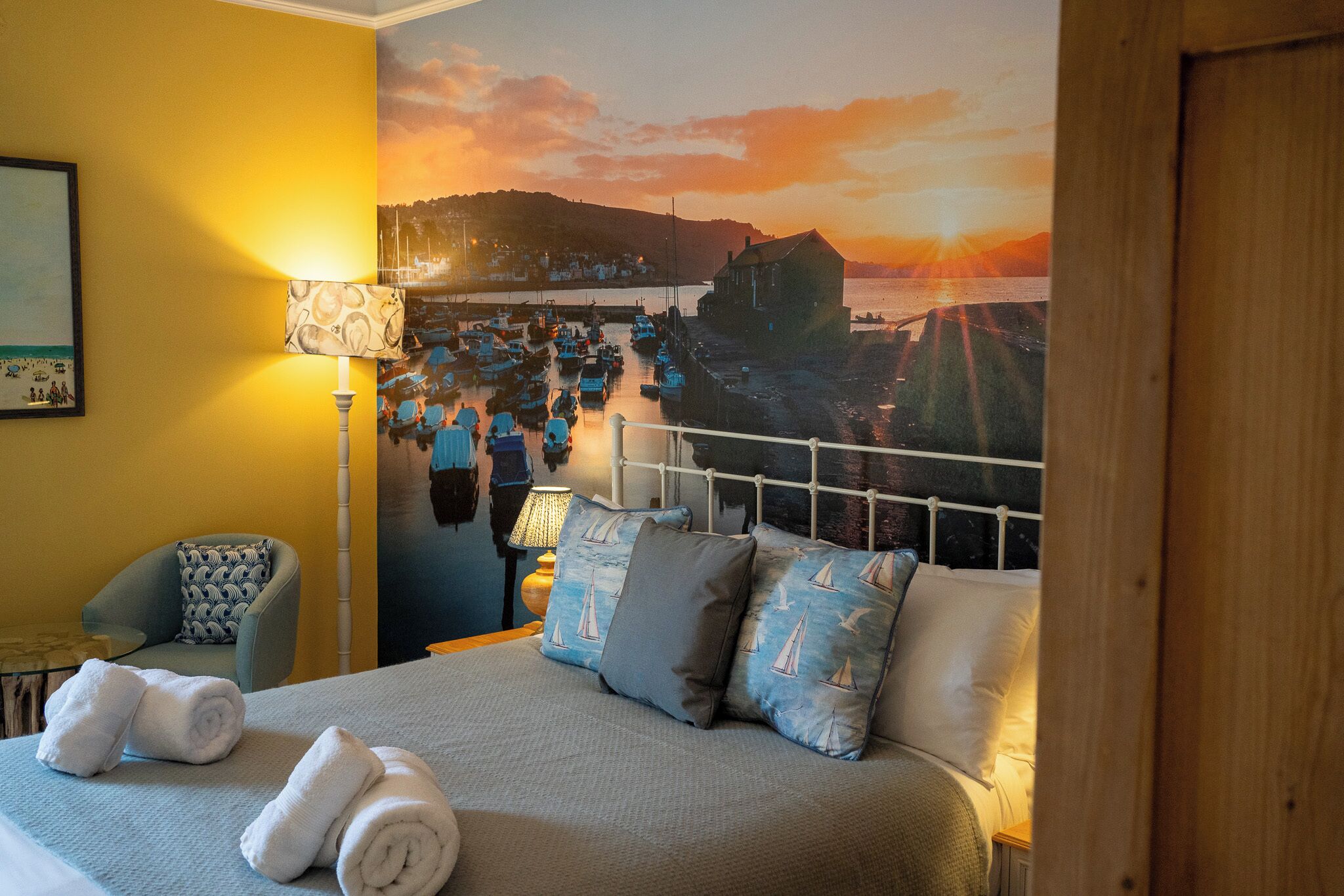 A hotel room with a bed, pillows, a chair, a lamp, and a mural of a harbor scene.