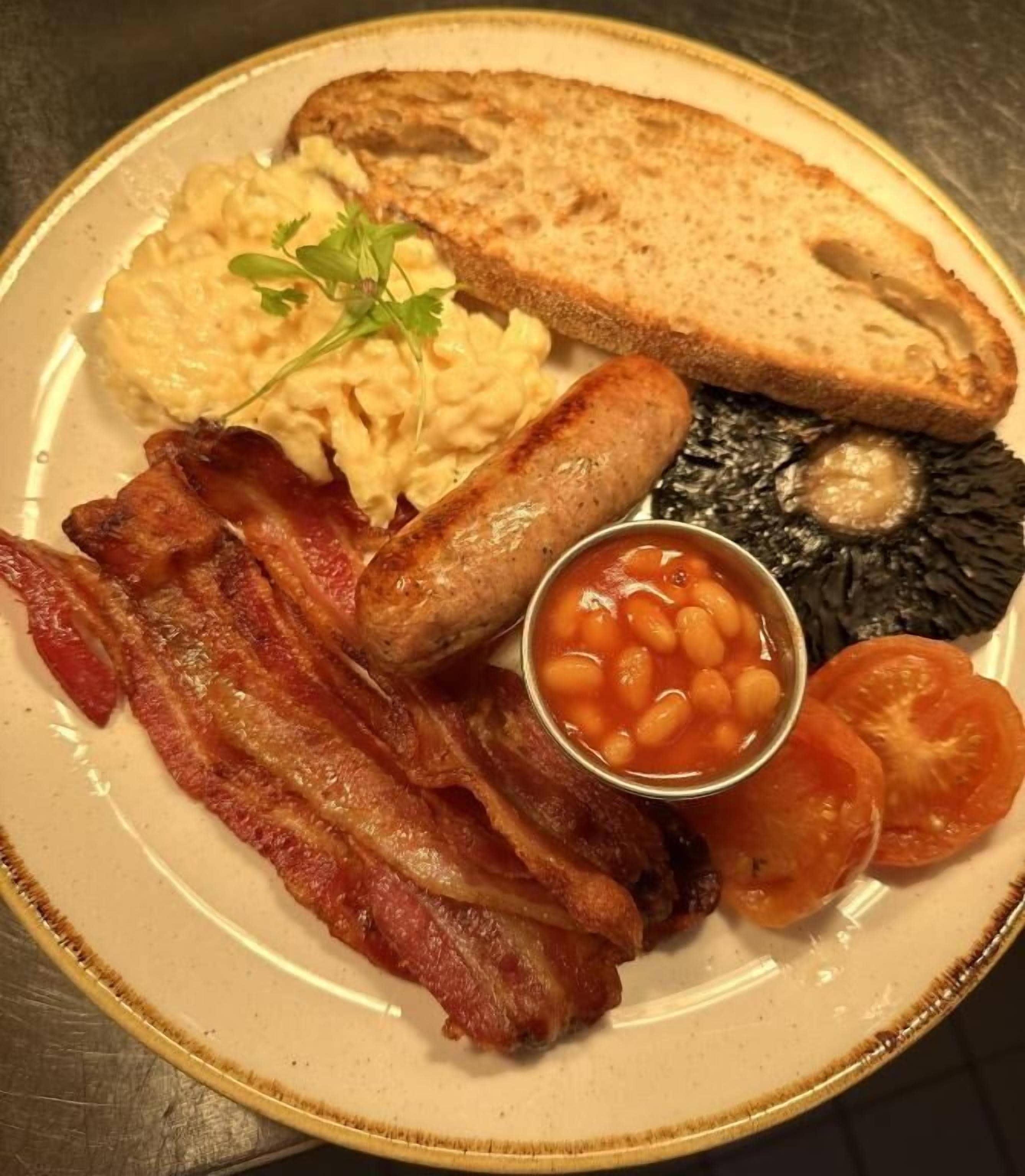 A plate with bacon, sausages, toast, scrambled eggs, baked beans, and tomatoes.
