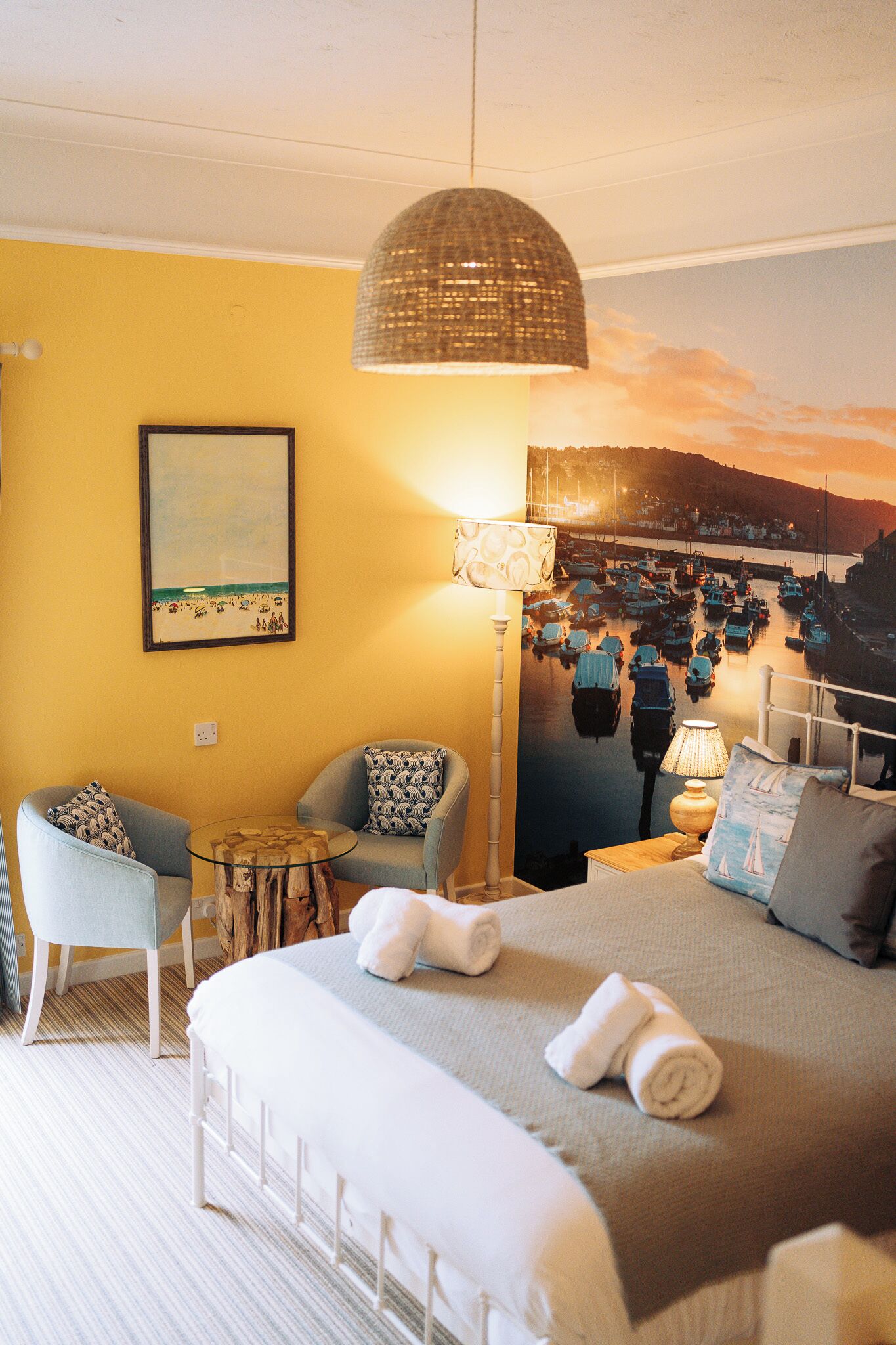 A hotel room with a bed, two armchairs, a small table, a lamp, and a painting of a harbor scene.