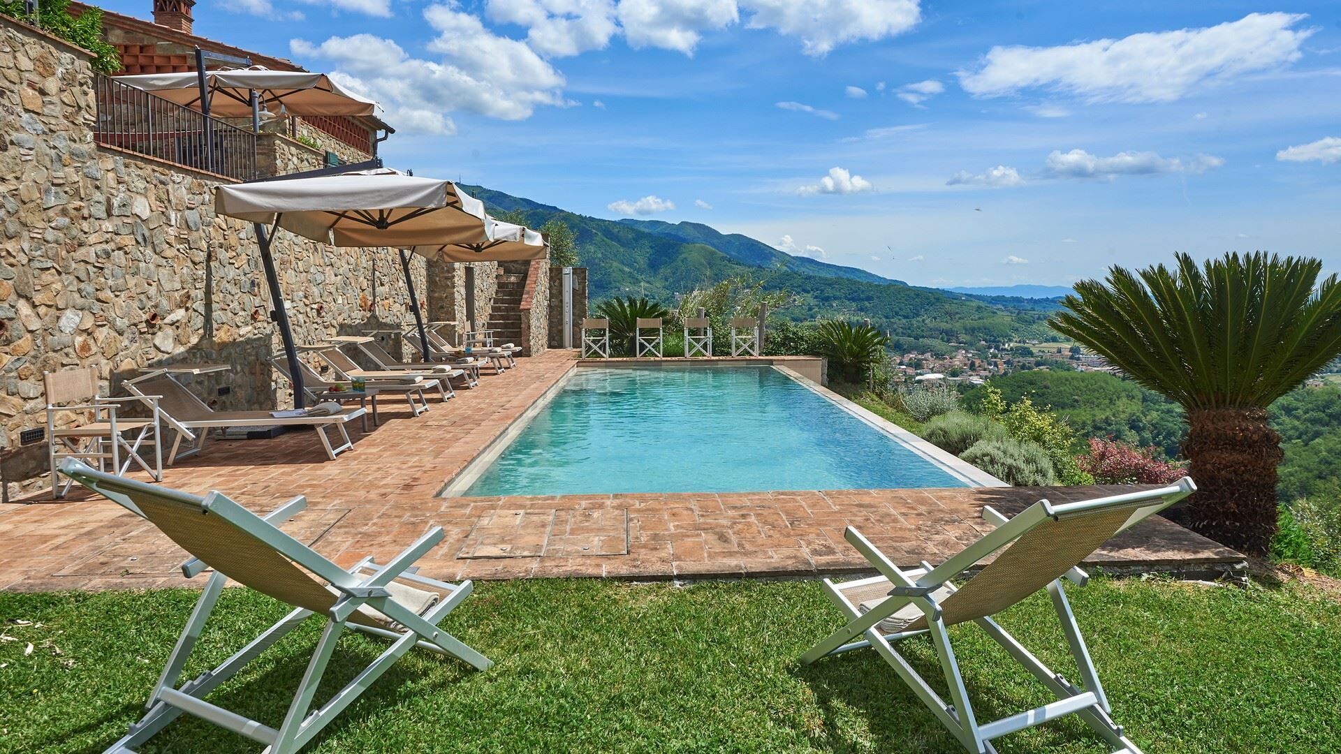 A pool with lounge chairs and umbrellas, surrounded by stone walls and a view of a town and mountains.