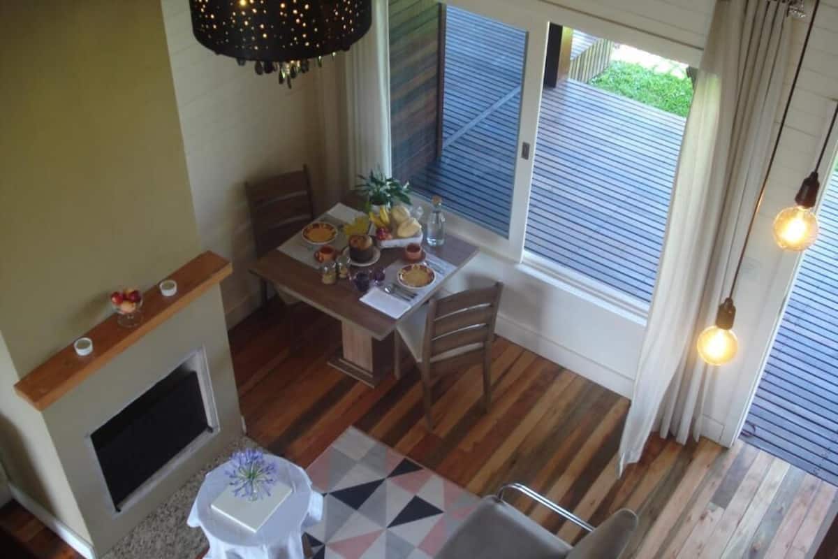 Uma sala de jantar com mesa de madeira posta para quatro pessoas, lareira e vista para o exterior através de grandes janelas.