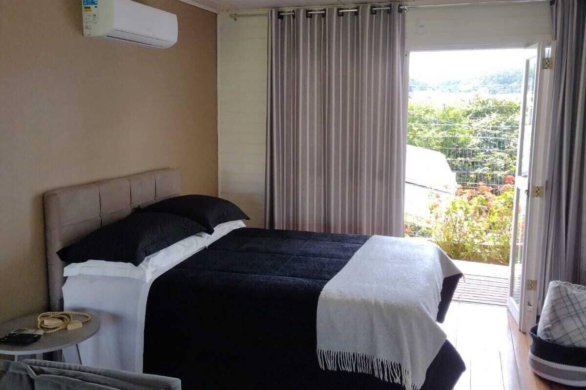 Quarto com cama, mesa de cabeceira, cadeira, janela com cortinas e vista para o verde lá fora.