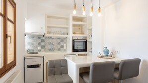 A modern kitchen with white cabinetry, a built-in oven, and a compact refrigerator. There are three bar stools and a dining table with a blue vase on it.