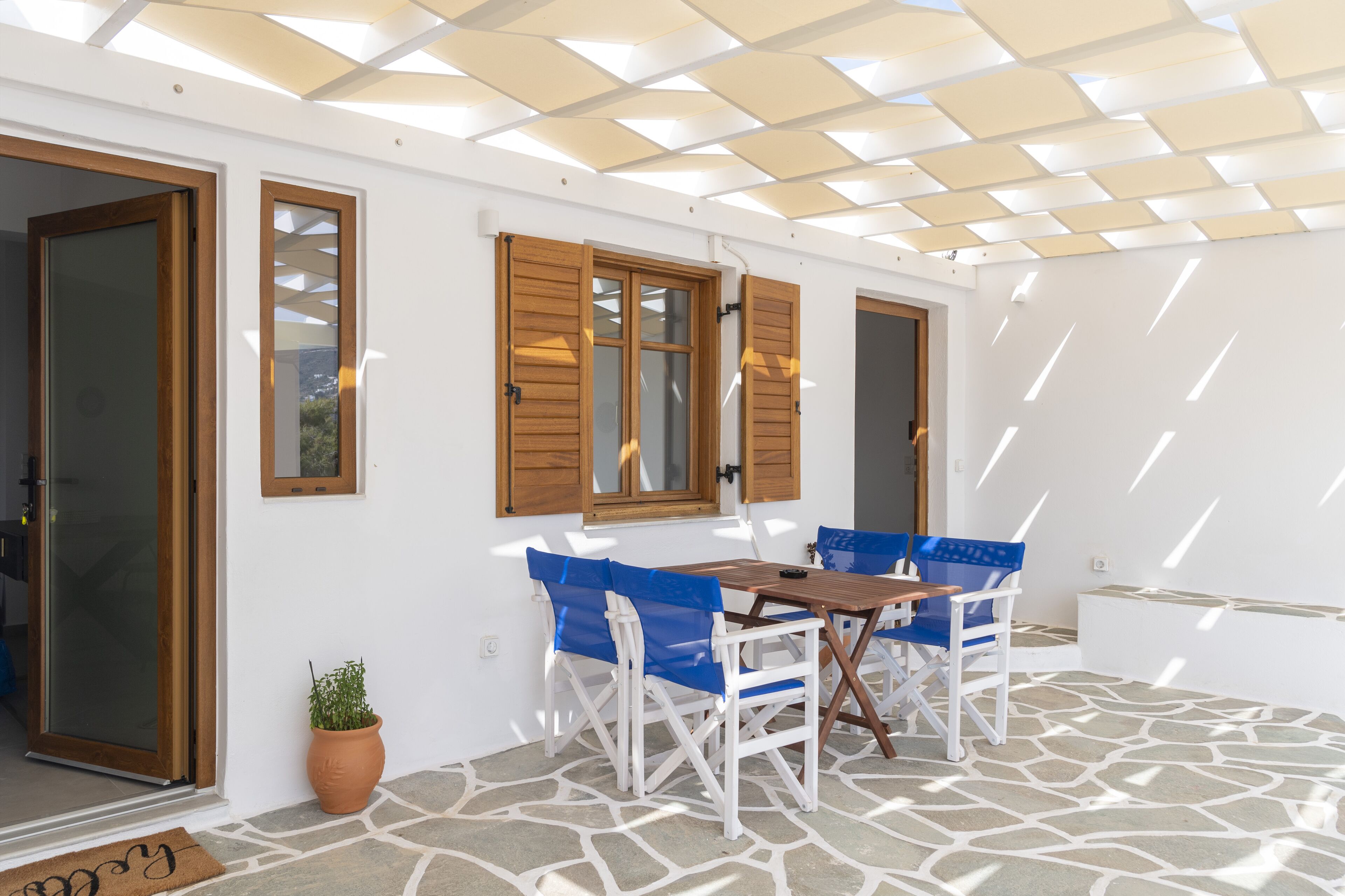 Un patio exterior con suelo de piedra, mobiliario de madera y una pérgola que da sombra.