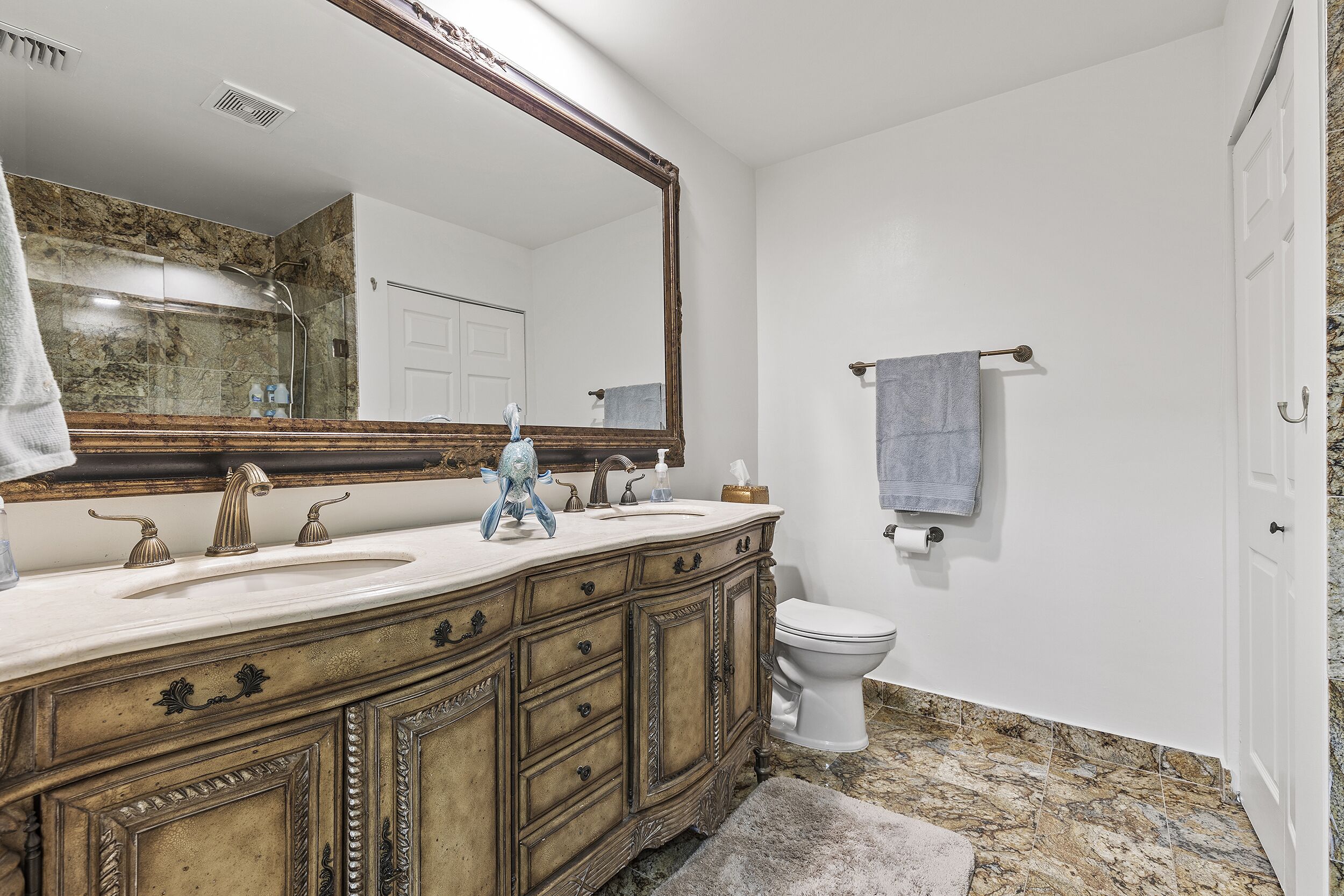Une salle de bain avec un sol en marbre, un grand miroir, des toilettes blanches et un meuble-lavabo en bois doté de tiroirs.