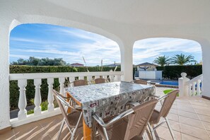 A covered outdoor patio with a table and chairs, a pool in the background, and a white railing.