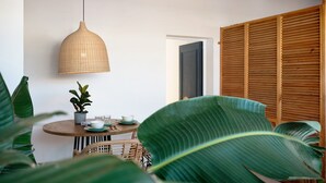 A round wooden table with a woven pendant light, a potted plant, and a door with wooden shutters.