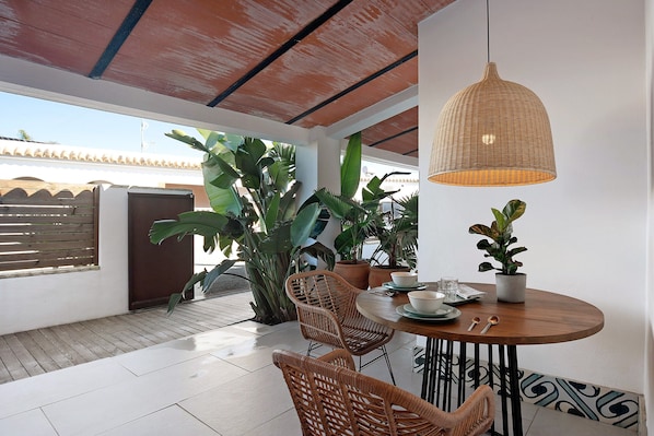A dining area with a round table, wicker chairs, a hanging light fixture, and potted plants.
