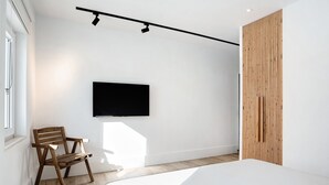 A modern bedroom with a bed, a wooden chair, a TV mounted on the wall, and a wooden panel on the right.
