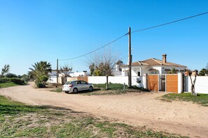 A residential area with a dirt road, a white car parked by the house, and a white building with a tiled roof.
