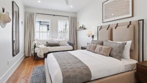 A bedroom with a large bed, a smaller bed, a ceiling fan, a window with curtains, a wall-mounted lamp, and a framed artwork.