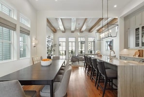 Uma sala de jantar espaçosa com uma mesa comprida, várias cadeiras e uma área de cozinha com bancada de mármore.
