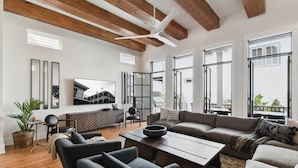 A spacious living room with a large sectional sofa, a coffee table, and a TV mounted on the wall. There are large windows providing natural light and a view of the outside. - Radiant New Construction | Heated Pool, Spa (New Orleans)