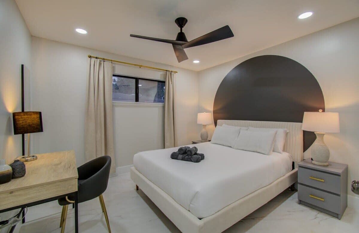 A modern hotel room with a bed, bedside lamps, a desk with a chair, a ceiling fan, and a window with curtains.