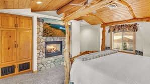 A bedroom with a stone fireplace, wooden ceiling beams, and a large bed. - Hot tub, ping pong, AC | 5 Suites (Sunriver)
