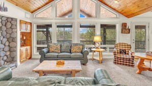 A spacious living room with a high wooden ceiling, a stone wall, a leather sofa, a wooden coffee table, and a view of the outdoors through large windows. - Hot tub, ping pong, AC | 5 Suites (Sunriver)
