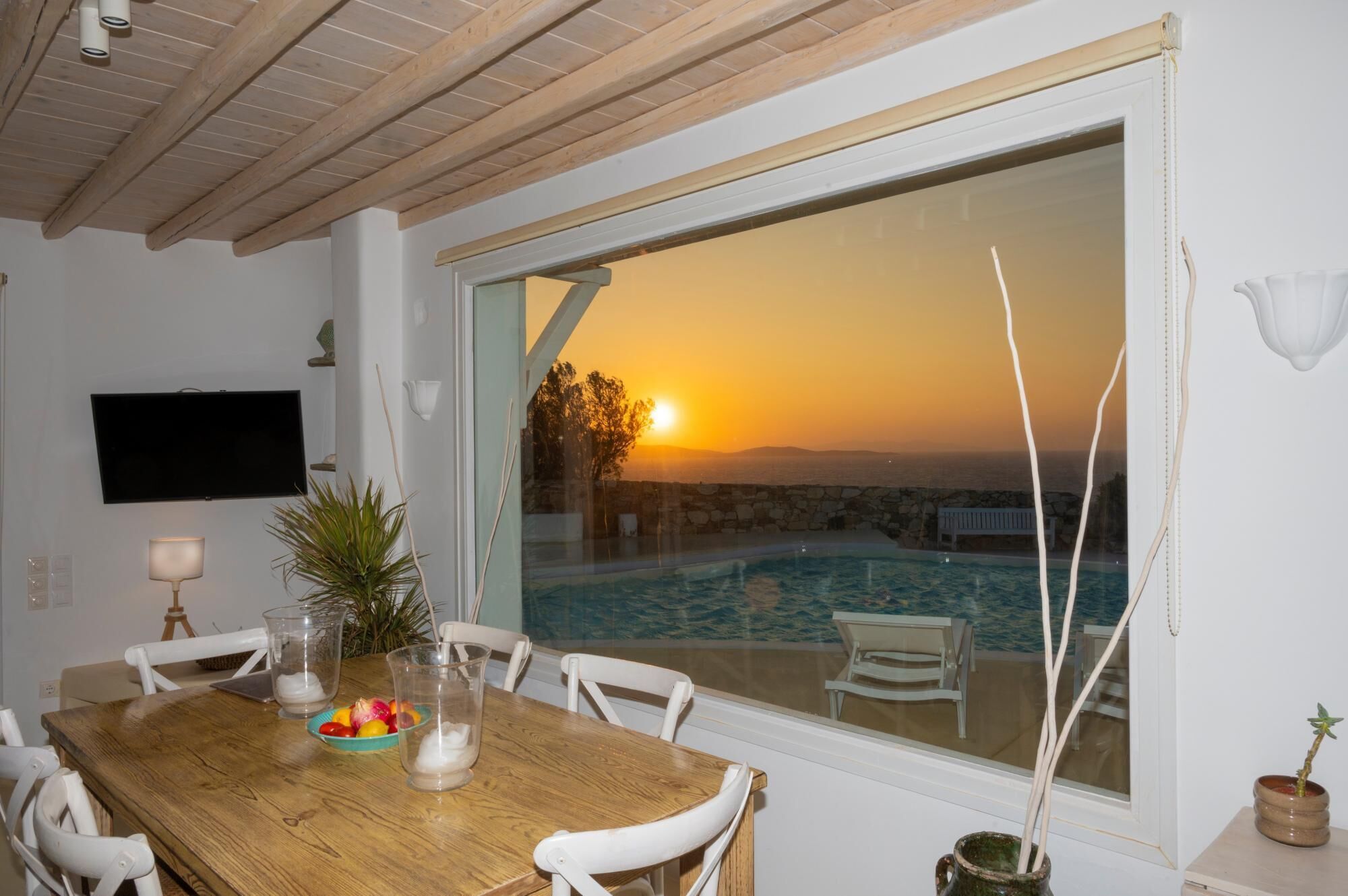Ein Essbereich mit Holztisch, Stühlen, einem Fernseher und Blick auf einen Pool bei Sonnenuntergang.