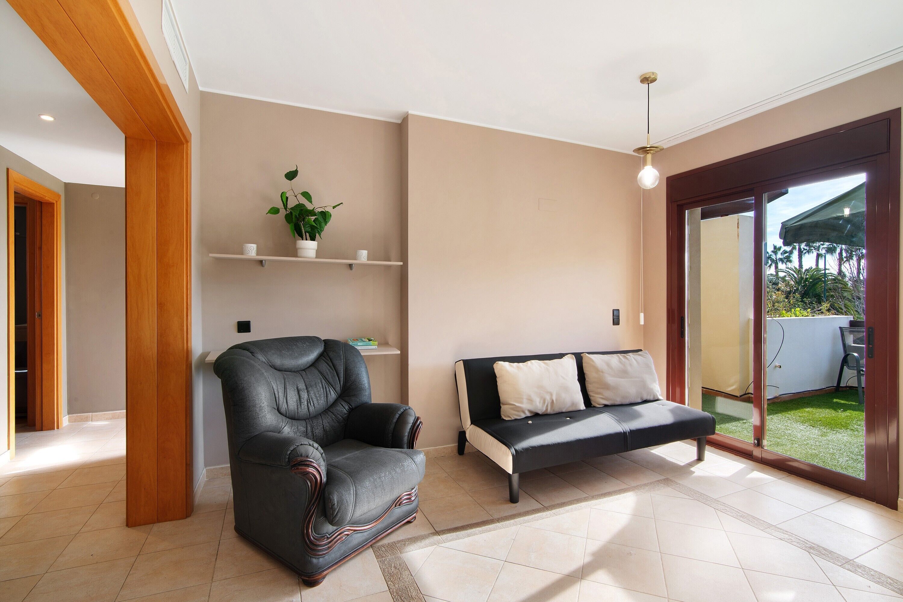 Una sala de estar con un sillón de cuero, un sofá, una planta en maceta y un ventanal corredero que da a un jardín.