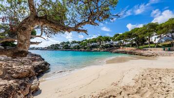 Ein Strand mit klarem türkisblauem Wasser, felsigem Ufer und weißen Gebäuden im Hintergrund.