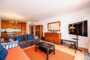 A living room with a sofa, coffee table, TV, wooden cabinets, and a kitchen area with appliances.
