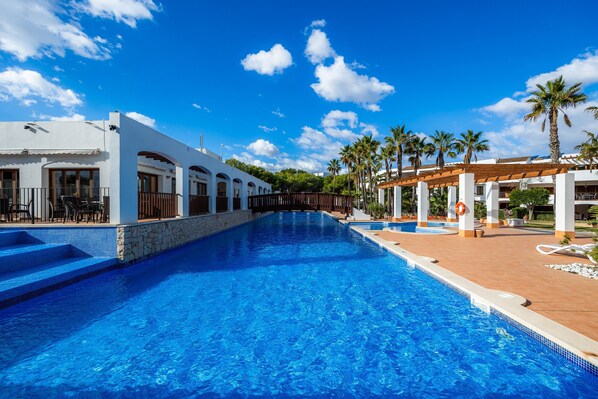 A swimming pool with steps leading into it, surrounded by white buildings with arched windows and a covered area with palm trees.