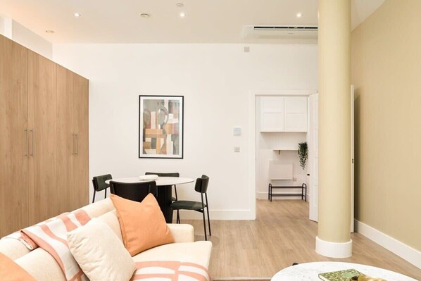 A modern living room with a sofa, a dining table with chairs, a kitchen area, and a framed artwork on the wall.