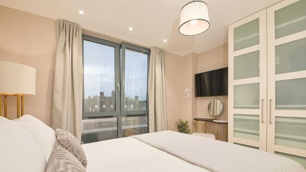 A hotel room with a large bed, a TV mounted on the wall, and a view of the city through the window. - Modern Marylebone apartment with balcony (London)