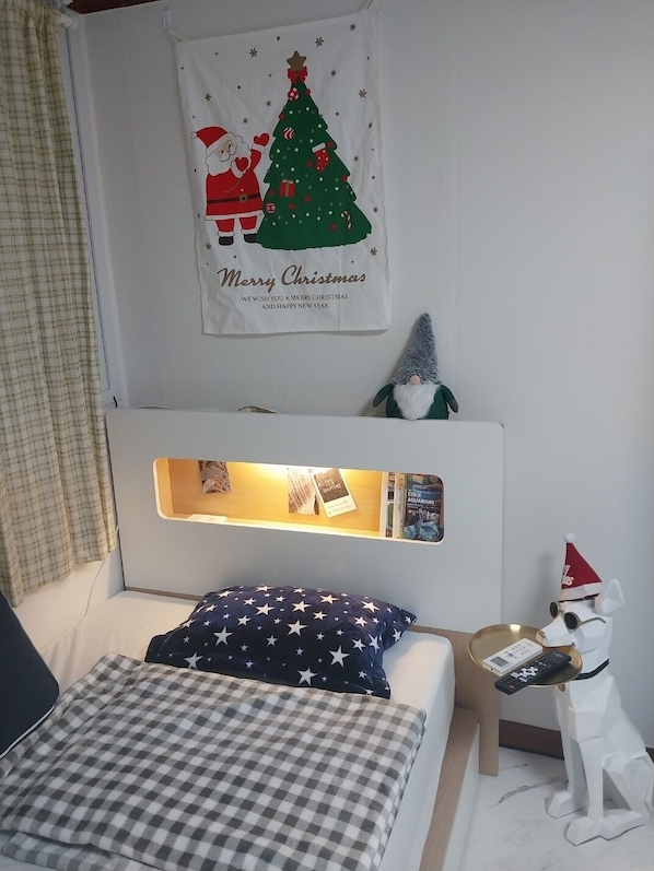 Une chambre à coucher avec un lit, une table de chevet, une étagère fixée au mur, une affiche sur le thème de Noël et une petite figurine de gnome.