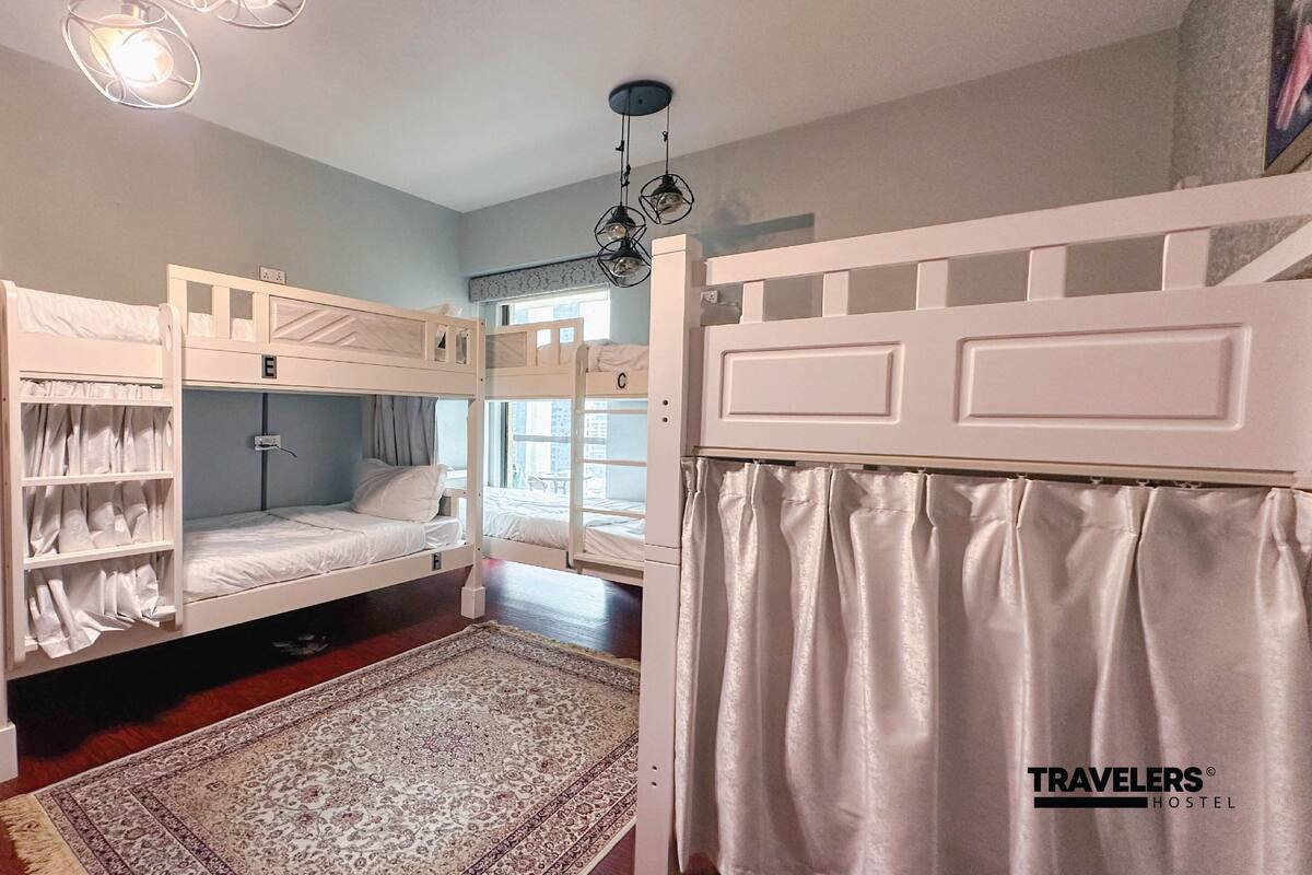 A dormitory room with bunk beds, a window with curtains, a patterned rug, and a ceiling light fixture.