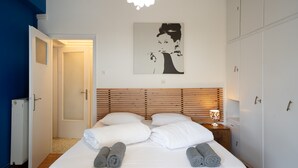 En pænt redt seng med hvide sengetøj og sammenrullede håndklæder, et træhovedgærde, et ophængt kunstværk og en sengetavlelampe.