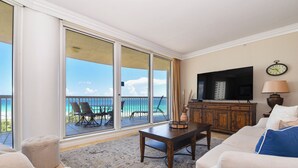 Ein Wohnzimmer mit einer großen Schiebeglastür, die auf einen Balkon mit Meerblick führt. Im Raum befinden sich ein Fernseher, ein Tisch und ein Sofa.
