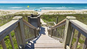 A wooden staircase leading to a beach with a clear view of the ocean and a flag. - Oceanfront home with stunning views, designed for comfort (Emerald Isle)