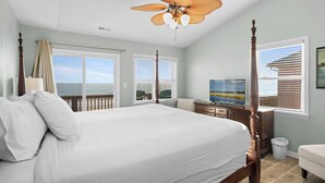 A bedroom with a large bed, a ceiling fan, a TV, and a view of the ocean. - Oceanfront home with stunning views, designed for comfort (Emerald Isle)