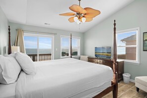 A bedroom with a large bed, a ceiling fan, a TV, and a view of the ocean.