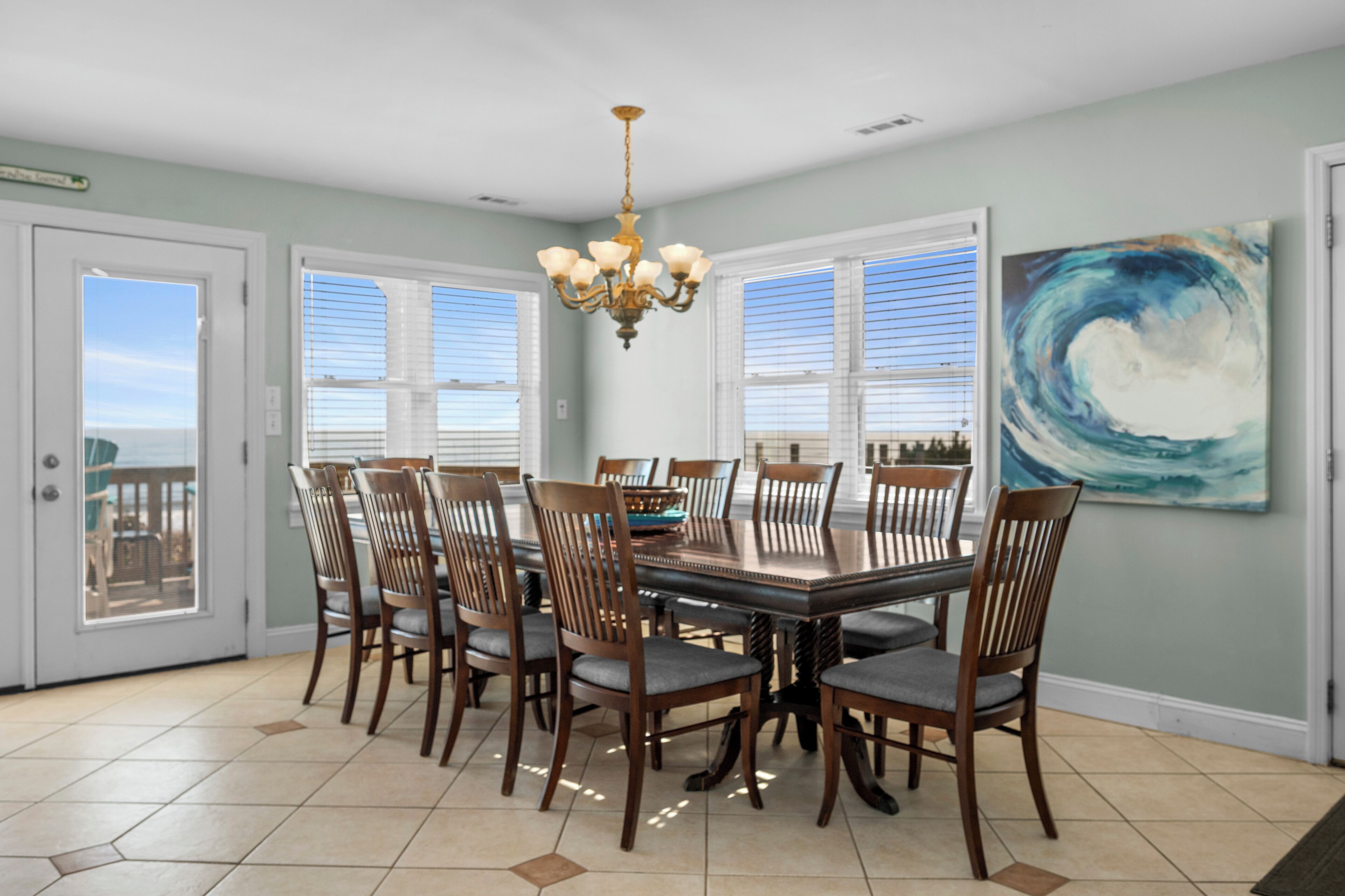 A dining room with a large table, six chairs, a chandelier, and a painting on the wall.