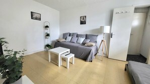 Un salon moderne et minimaliste, avec un canapé gris, une petite table blanche et un lampadaire. Deux tableaux encadrés ornent le mur, et une plante en pot se trouve sur la gauche.