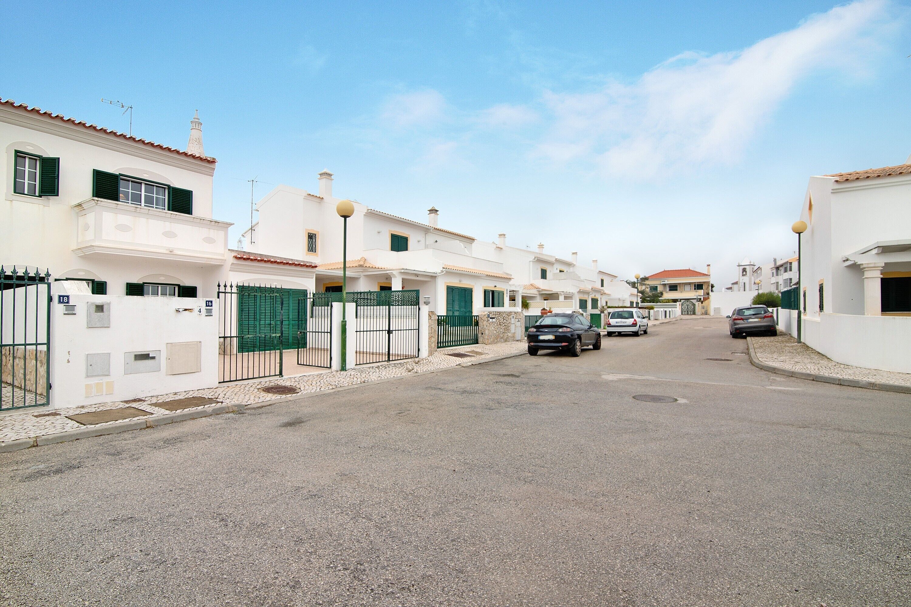 A residential street with white houses, green gates, and parked cars.