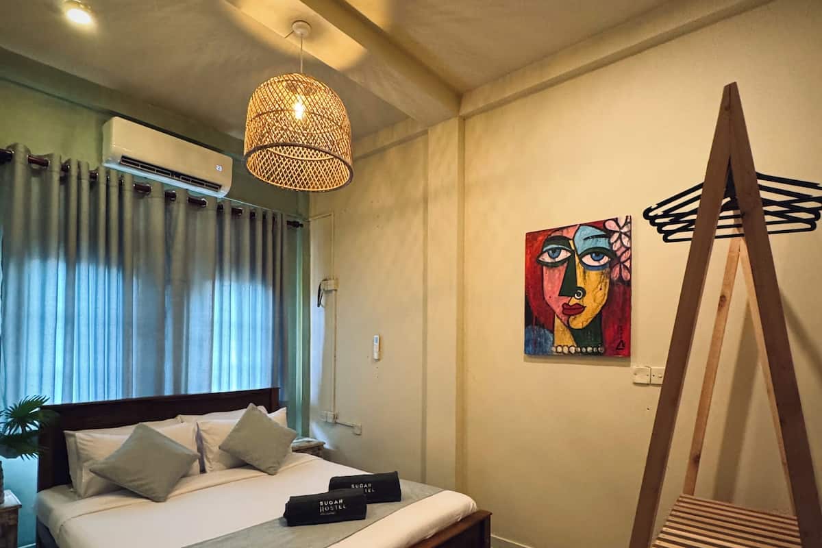 Ein Schlafzimmer mit einem Bett, einer Hängeleuchte, einer Klimaanlage, einem Gemälde und einem Holzstuhl.
