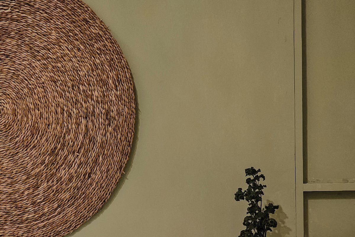 Ein Bett mit weißen Leinen und einem grünen Kopfkissen, ein gewebter Wandbehang und ein Blumentopf auf einem Nachttisch aus Holz.