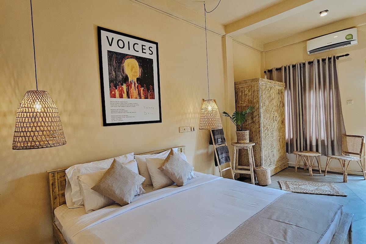 Ein Hotelzimmer mit einem großen Bett, zwei Pendelleuchten, einem Poster an der Wand, einem Schreibtisch mit Stuhl und einem Fenster mit Vorhängen.