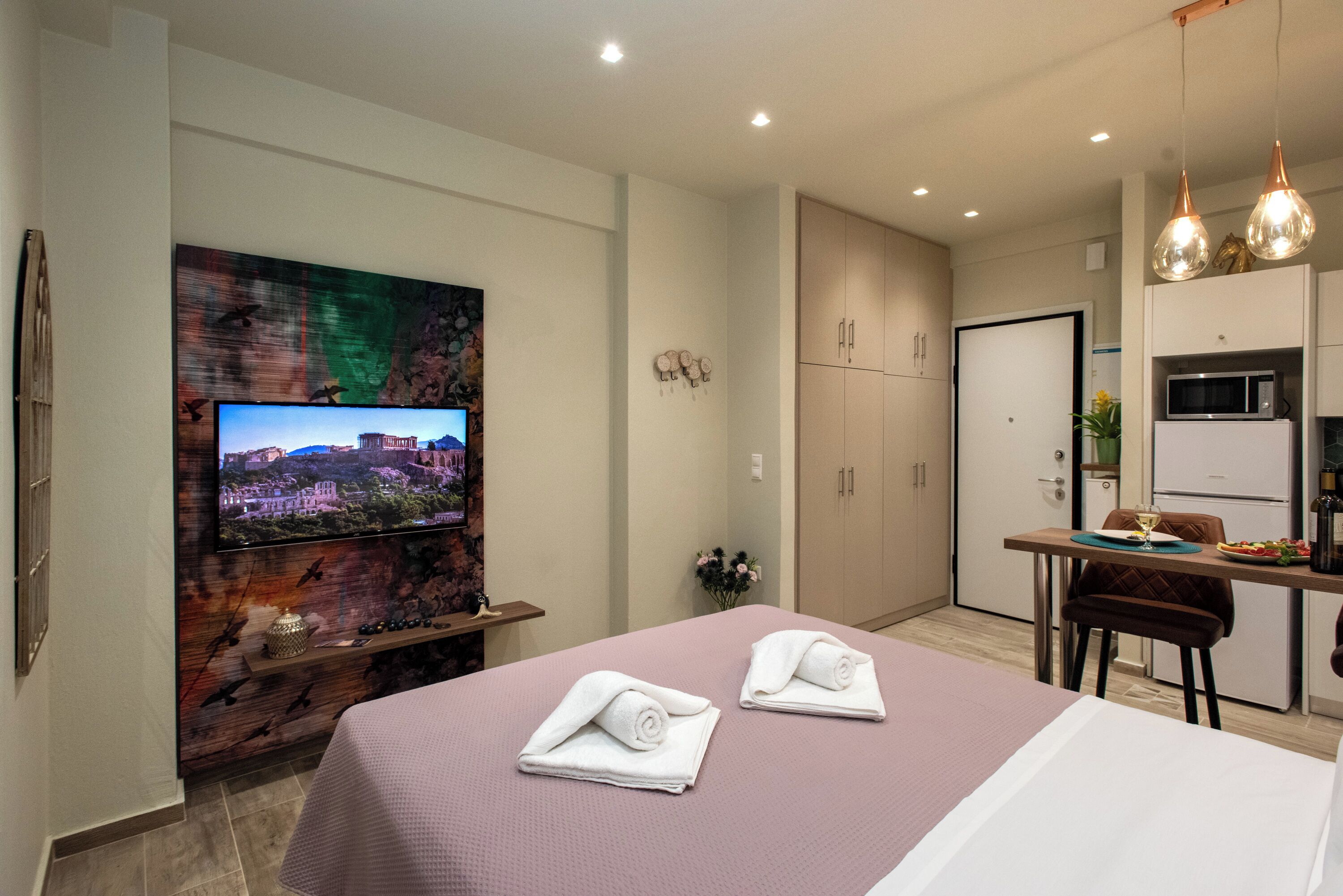 A modern hotel room with a bed, a TV mounted on a wall, a small kitchen area, and a dining table.