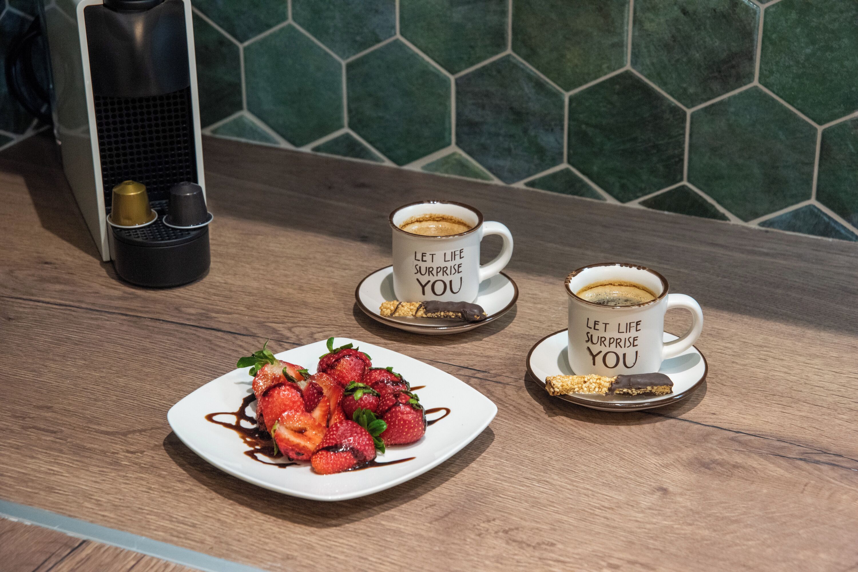 A breakfast setting with two coffee cups on saucers, a plate of strawberries with chocolate sauce, and a coffee machine.