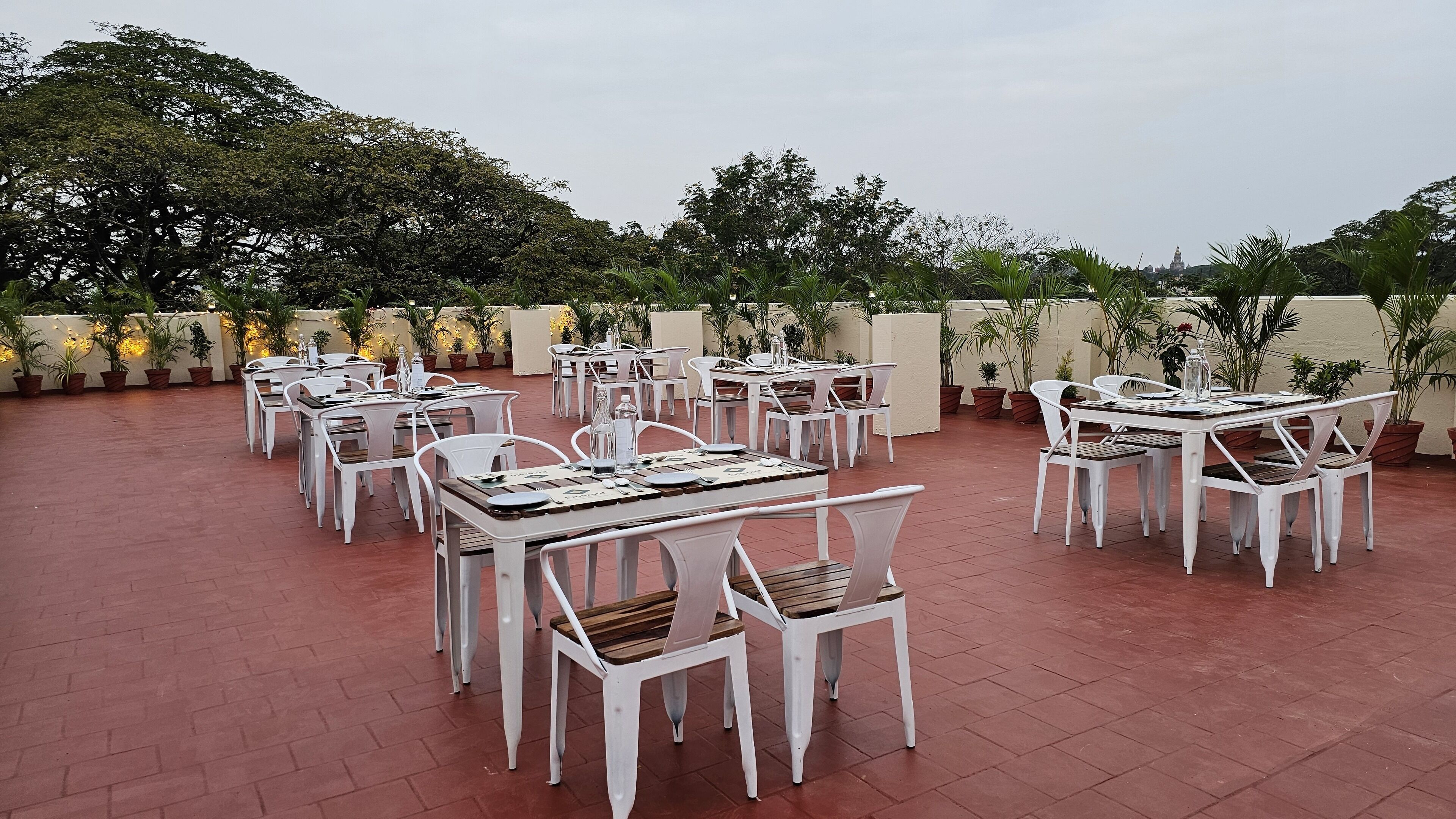 An outdoor patio with white tables and chairs, surrounded by potted plants and trees.