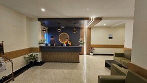Uma área de recepção de hotel com balcão, dois recepcionistas, sala de espera com cadeiras e um vaso de planta.